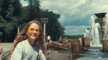 Uzun saçlı ve gözlüklü bir kız çeşmenin etrafında dolanıyor..