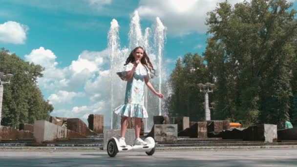En flicka i en lång klänning rider en gyro scooter nära fontänen. — Stockvideo
