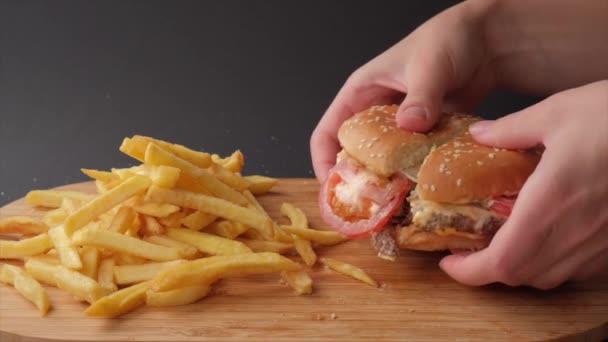 Rozerwij hamburgera na pół rękami. Na stole są frytki i hamburger.. — Wideo stockowe