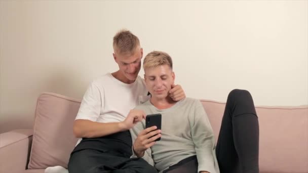 Pasangan gay memeluk dan berbaring di sofa, memegang ponsel. — Stok Video