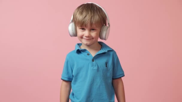 Menino bonito em fones de ouvido brancos ouve música e olha para a câmera. — Vídeo de Stock