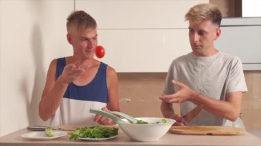 Genç sarışın adam bir arkadaşına domates fırlatıyor. Salata için sebze doğra..