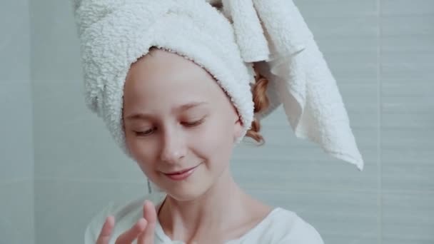 Een jong meisje met een witte handdoek op haar hoofd brengt een vochtinbrengende crème op haar gezicht. — Stockvideo