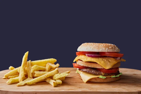 Vedle hranolek na lehkém dřevěném stole sedí velký hamburger. Tmavé pozadí. — Stock fotografie