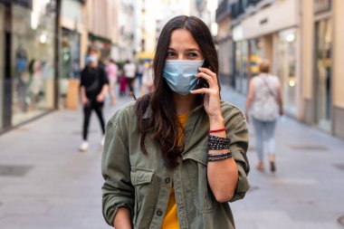 Sokakta cep telefonu kullanırken yüzüne koruyucu maske takan bir kadın.