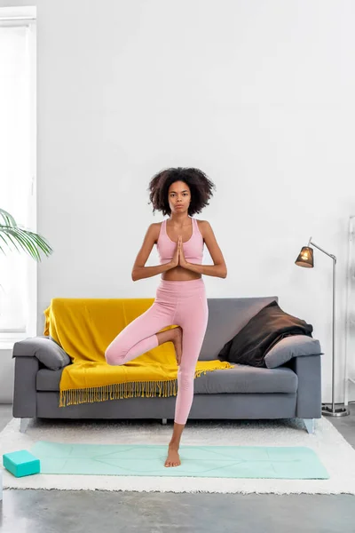 Afroamerican woman practicing yoga tree pose at cozy home