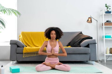 Afro kadın yoga yapıyor. Nilüfer çiçeği pozu veriyor. Evinde meditasyon yapıyor.