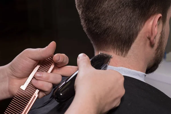Hairdresser cuts hair with a trimmer for a young guy with a beard. Close-up of a master haircutting male hair. Cutting hair at the temples of a guy. Bearded man at the hairdresser. Men\'s haircut
