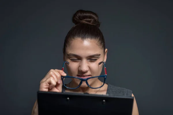 Strict young woman holding large nerd glasses Portrait of a squinting girl, staring hard at a notebook, not seeing letters. Studio photo on a gray background. Poor eyesight. Visual impairment due to vitamin A deficiency. It is time to see an ophthalm