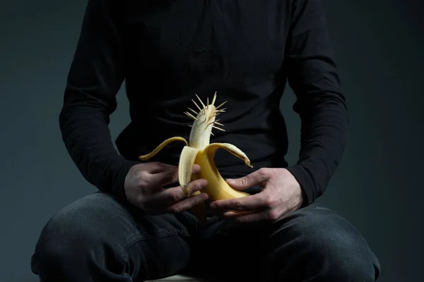 photo of a young man fighting against erection problem concept of genital cancer in men. A man in black holds a banana in his hands with thorns on his head, with a background on the male penis. On a gray background. Male genital diseases. Prostate ca