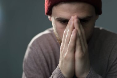 Genç sakallı bir adamın portresi, elleri yüzüne yapışık, dua ediyor, gözlerini aşağı indiriyor. Gri arka planda stüdyo fotoğrafı. 25 yaşında üzgün bir genç adam, üzüntü, özlem, depresyon, yardıma ihtiyacı var, uyuşturucu bağımlısı, hüküm giymiş, otizm, p