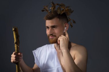 Kablosuz kulaklıklarla antik Yunan sakallı tanrısı, bir asayla defne çelenginde müzik dinliyor. Stüdyo fotoğrafı dramatik bir şekilde, gri bir arkaplanda. Beyaz çarşaflı Olimpiyat Tanrısı, elinde bir sopayla, kablosuz kulaklıkla müzik dinliyor. Karşılaştır