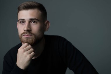 25 yaşında düşünceli genç bir adam. Eli çenesine dayanmış, siyahlar içinde düşünen sakallı bir adam. Gri arka planda stüdyo fotoğrafı. Bir filozof, düşünür, bir adam düşünür, bir bakış, bir insan portresi.