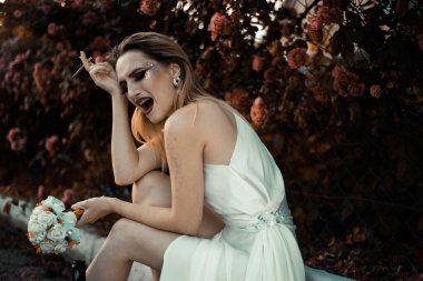 Beyaz elbiseli gelin ağlıyor, elinde bir sigara, elinde bir şişe alkol tutuyor. Kırmızı güllerin çiçekli arka planına karşı