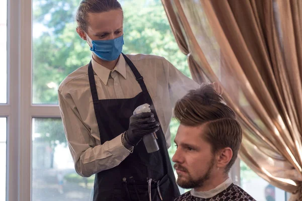 Um barbeiro com uma máscara protectora numa barbearia está a cortar um jovem barbudo. corte de cabelo de quarentena, trabalhador da barbearia com uma máscara protetora a rapar um tipo — Fotografia de Stock