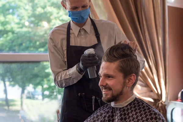 En frisör i skyddsmask i en frisörsalong skär en ung skäggig kille. karantänfrisyr, frisör arbetare i en skyddande mask rakning en kille — Stockfoto