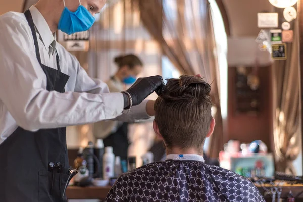 En frisör i skyddsmask i en frisörsalong skär en ung skäggig kille. karantänfrisyr, frisör arbetare i en skyddande mask rakning en kille — Stockfoto