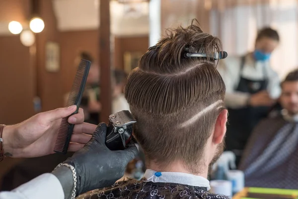 a hairdresser cuts a bearded young guy with a hair trimmer, combing the hair on his head. Work of the master in mens haircut in a barbershop