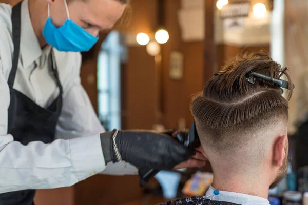 En frisör klipper en skäggig ung kille med en hårtrimmer, kammar håret på huvudet. Mästarens arbete i herrfrisyr i en frisörsalong — Stockfoto