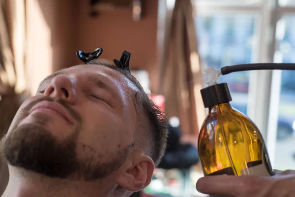 En hårfrisör som sprejar ett balsam efter att ha rakat ett skägg till en kund till en ung skäggig kille som är 25 år gammal. Till frisersalongen — Stockfoto