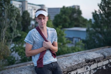 Elleri göğsünde duran kırmızı kazaklı, beyaz beysbol şapkalı bir gencin portresi.