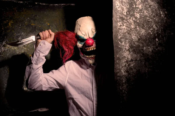 scary clown, in the shadows, looking eerily at the camera from behind an abstract wall. Halloween concept. A very scary clown with a knife in his hand, looks menacingly at the camera, against a dark abstract background. Halloween clown, killer, horro