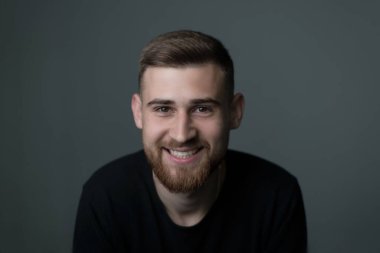 Genç sakallı, neşeli bir adamın portresi, 25 yaşında, siyahlar içinde. Kameraya bakıyor, gülümsüyor. Gri arka planda stüdyo fotoğrafı. Neşe, kahkaha, sakallı bir adamın gülümsemesi, genç adam