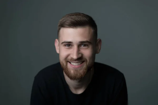 portrait of a young bearded cheerful guy, twenty-five years old, in black. looking at the camera, smiling. Studio photo on a gray background. Joy, laughter, smile of a bearded man, young guy