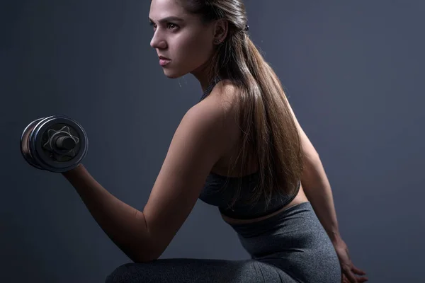 Brutal athletic woman pumping up muscles with dumbbells portrait of sportswoman doing exercise with dumbbell. Studio photo on a gray background. Sport and health. Active lifestyle. Athletic girl shakes the muscles of the hands of a dumbbell.