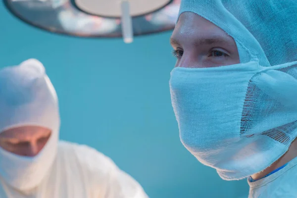 close-up of surgeons in a sterile mask, against the background of an operating lamp, with raised hands, the surgeon\'s assistant looks at the indicators in a monitor, sterile gloves and a mask. Pediatric surgery in Ukraine. Sterile conditions, medical