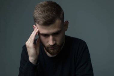 Baş ağrısından muzdarip 25 yaşlarında, siyah giysili genç bir adam elini kafasına bastırıyor ve baş ağrısı çekiyor. Gri bir arka planda. Depresyonda, sağlıksız bir adam, baş ağrısı, migren, akıl sağlığı, üzüntü, zayıflık, baş dönmesi