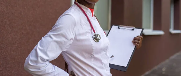 African American Nurse black young woman doctor, in a white coat, with a phonendoscope, holds a folder for medical papers in a rue, against the background of a hospital. A young black girl of twenty seven years old, a doctor in a white coat, smiles,