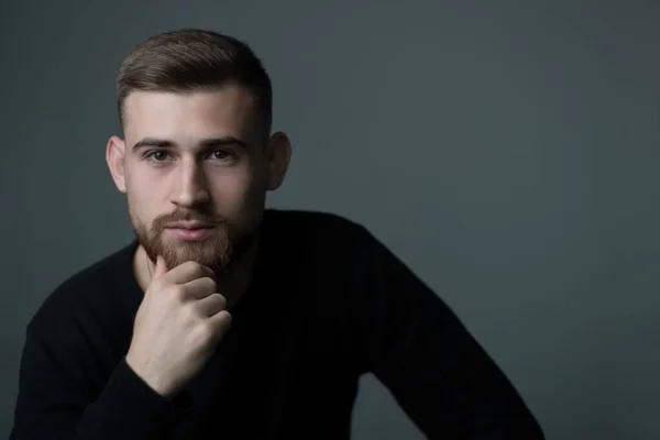 pensive young man of twenty five years old. A bearded man thinking, holding his hand to his chin, in black. Studio photo on a gray background. A philosopher, thinking, a man thinks, a look, a portrait of a man.