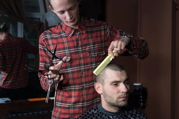 hairdresser a young guy with many scissors in his hand, cuts with a grimace a young guy client, in a barbershop. Hair care, young men of twenty-five years old in a barbershop, a hairdresser cuts the holder in his hand with the help of many scissors