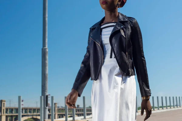 African fashion model, in black leather jacket, white dress, posing. Against the background of street texture. Young black girl, twenty-seven years old, in a black leather jacket, white and pink dress, posing, street fashion