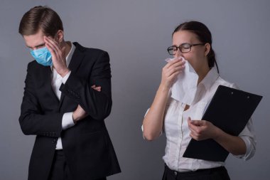 İş elbiseli iki genç iş adamının portresi. Maskeli adam hasta bir kızın enfeksiyonundan korkarak yüzünü eliyle kapatıyor. Mendilin içine hapşırıyor. Hasta bir ofis çalışanının iş anlayışı. Grip salgını. Gri bir B 'de stüdyo fotoğrafı.