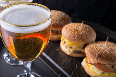 Hamburger ve patatesli bir kupa bira, el yapımı koyu ve köpüklü bira bardakları, hamburgerlerin arka planında, Shyomga, sığır eti köftesi, peynirli burger gibi çeşitli malzemelerle. Köy tarzında, bir siyaha karşı.