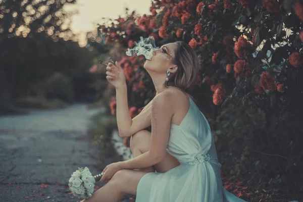 Traurige Braut raucht eine Zigarette und trinkt Alkohol. Vor dem Hintergrund eines floralen Hintergrunds aus Rosen. Traurige Braut beim Zigarettenrauchen, mit einer Flasche Alkohol in der Hand — Stockfoto