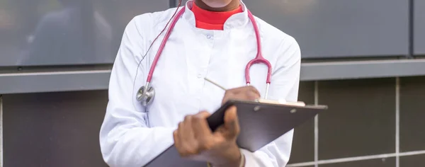 Афро-американський лікар, якому 27 років, в білому пальто, з фонендоскопом, пише ручку в папку для паперів. На чорному тлі. — стокове фото