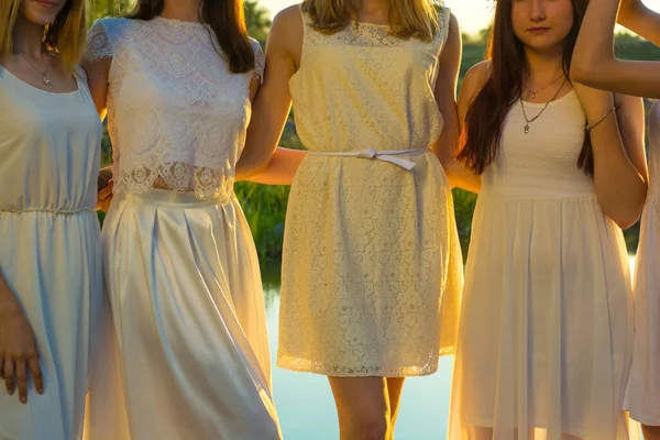 Μια ομάδα νεαρών κοριτσιών 20 ετών, με λευκά φορέματα, στεφάνια λουλουδιών, αγκαλιασμένα, να στέκονται στο ηλιοβασίλεμα στη λίμνη. Εθνική εορτή των νύφων — Φωτογραφία Αρχείου