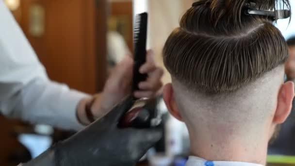 Un coiffeur coupe un jeune homme barbu avec une tondeuse à cheveux, peignant les cheveux sur sa tête. Travail du maître en coupe de cheveux pour hommes dans un salon de coiffure — Video