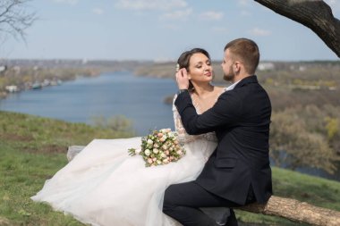 Damadın genç çifti, gelinle öpüşüyor, sarılıyor, doğal bir arka planda. Yaz düğünü. Sevecen bir çift, bir düğün töreni, 25 yaşında yeni evli bir çift, doğal bir geçmişe karşı, bir bankta oturuyorlar.