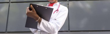 Afro-Amerikalı kadın doktor, 27 yaşında, beyaz önlüklü, fonendoskoplu, kağıtlar için bir klasöre kalem yazıyor. Siyah bir arka planda. Tıbbi geçmişi. Tıbbi konsept, siyah tenli genç doktor, elinde bir klasör, sağlık hizmeti.