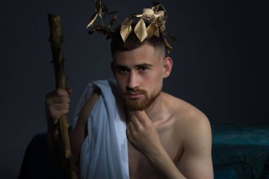 Beyaz elbiseli, başında çelenk olan antik Yunan tanrısı Olympus 'un portresi, siyah bir bankta oturan bir asa tutuyor. Gri bir arka planda. Bereket tanrısı, Asclepius, 25 yaşında genç bir adamın portresi.