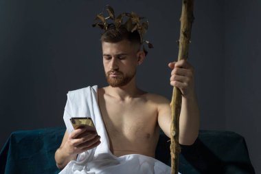 Beyaz çarşaflı, gri bir arka planda akıllı telefonuyla konuşan Olimpik sakallı genç tanrı. Antik Yunan ve bugün çağının konsepti. Modern teknoloji cep telefonu, akıllı telefon, Tanrı akıllı telefonla konuşuyor.