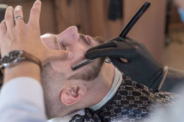 Berber, genç sakallı bir berber dükkanını tıraş ediyor. Kapat. Sakal kesimi, tehlikeli jilet, profesyonel saç ve sakal bakımı. Berber dükkanında tıraş olan bir adam..