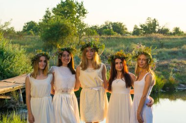 20 yaşında bir grup genç kız, beyaz elbiseli, çiçeklerden çelenkler takmış, sarılıyorlar, gün batımına karşı duruyorlar. Ulusal gelin bayramı, çiçeklerin başında çelenkler, genç kızlar, bekarlık konsepti, bekarlığa veda partisi.