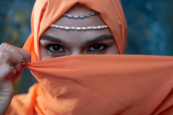 close-up portrait of a young Arab girl with beautiful eyes in an orange burqa, holding her hands near her face, on a dark background. She hid her face in a handkerchief. The ritual of tying the burqa. The girl hid her hair behind an orange silk cloth