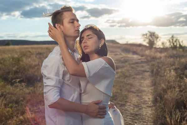 portrait of a young wedding couple, a guy and a girl of twenty-two years old, in a field with a flying white ribbon behind, against the backdrop of the setting sun, cloudy blue sky. Amorous couple, look passionately at each other, white wedding dress