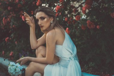 Beyaz elbiseli üzgün bir gelinin sanat portresi, elinde bir sigara, bir şişe alkol tutuyor. Kırmızı gül dokusunun arka planında, doğal çiçek arkaplanı. Üzgün gelin, gelinlik içinde sigara içmenin zararı. Yaratıcı sanat 